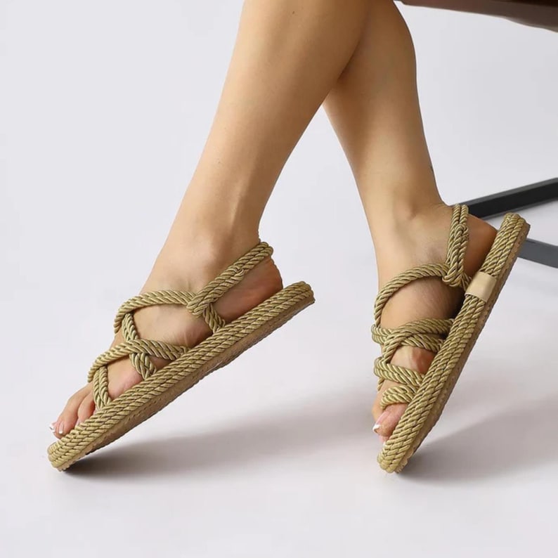 May include: Tan rope sandals with a woven design. The sandals feature a flat sole and straps that wrap around the foot and ankle. The sandals are made of a natural-looking material and are suitable for casual wear.