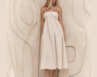 May include: A cream-colored midi dress with a halter neckline and a slit in the front. The dress has a fitted bodice and a flowing skirt. The model is wearing clear heels. The background is a light-colored wall with a textured design.