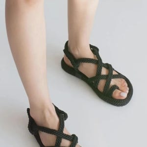 May include: Black rope sandals with a woven design. The sandals have a flat sole and a strap that goes around the ankle. The sandals are shown on a white background.