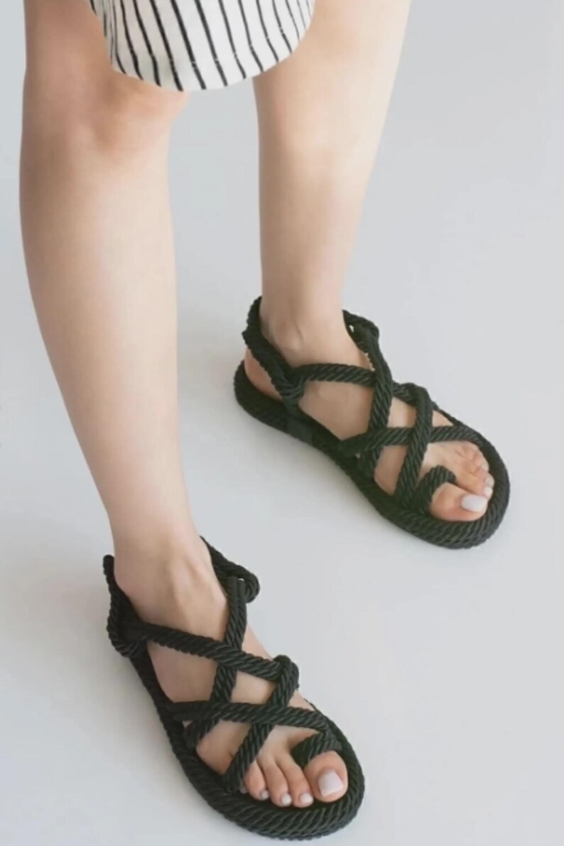 May include: Black rope sandals with a woven design. The sandals have a flat sole and a strap that goes around the ankle. The sandals are shown on a white background.