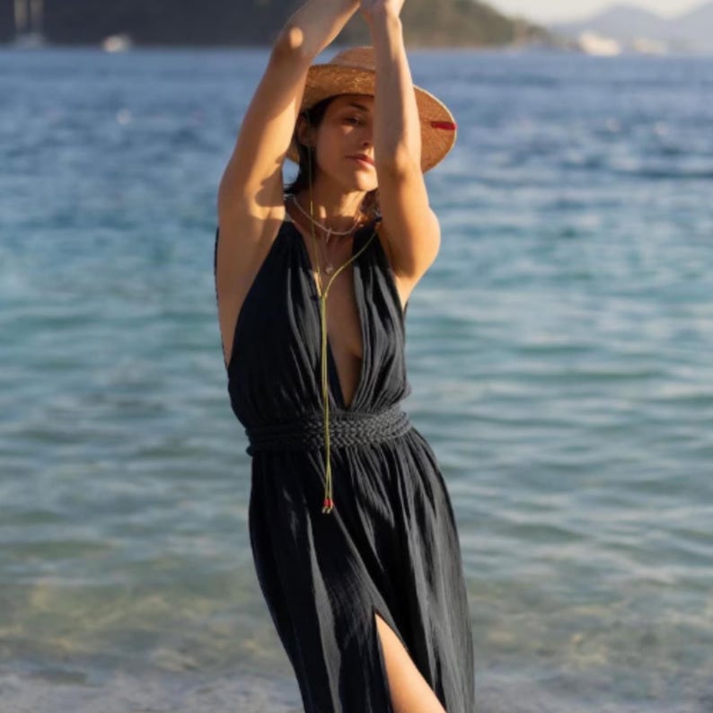May include: A woman wearing a dark blue maxi dress with a plunging neckline and a braided belt. She is wearing a straw hat with a red band and a gold chain necklace. The woman is standing on a beach with the ocean in the background.