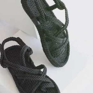 May include: A pair of black rope sandals. The sandals feature a woven design on the footbed and straps that wrap around the ankle and foot. The sandals are displayed on a white surface.