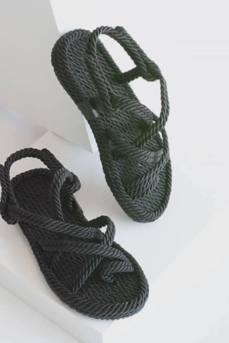 May include: A pair of black rope sandals. The sandals feature a woven design on the footbed and straps that wrap around the ankle and foot. The sandals are displayed on a white surface.