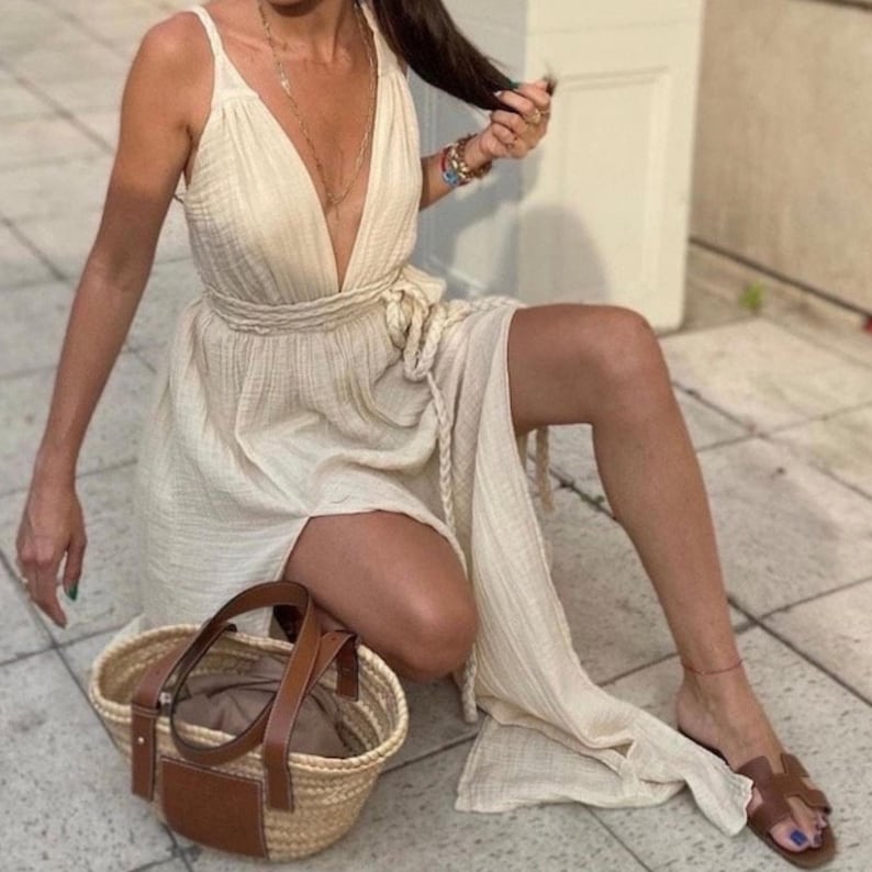 May include: A woman wearing a white maxi dress with a deep V-neckline and a side slit. She is sitting on a stone floor with a brown leather and straw tote bag beside her.