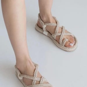 May include: Beige rope sandals with a toe loop and crisscross straps. The sandals have a flat sole and a strap around the ankle. The footwear is displayed on a white background.