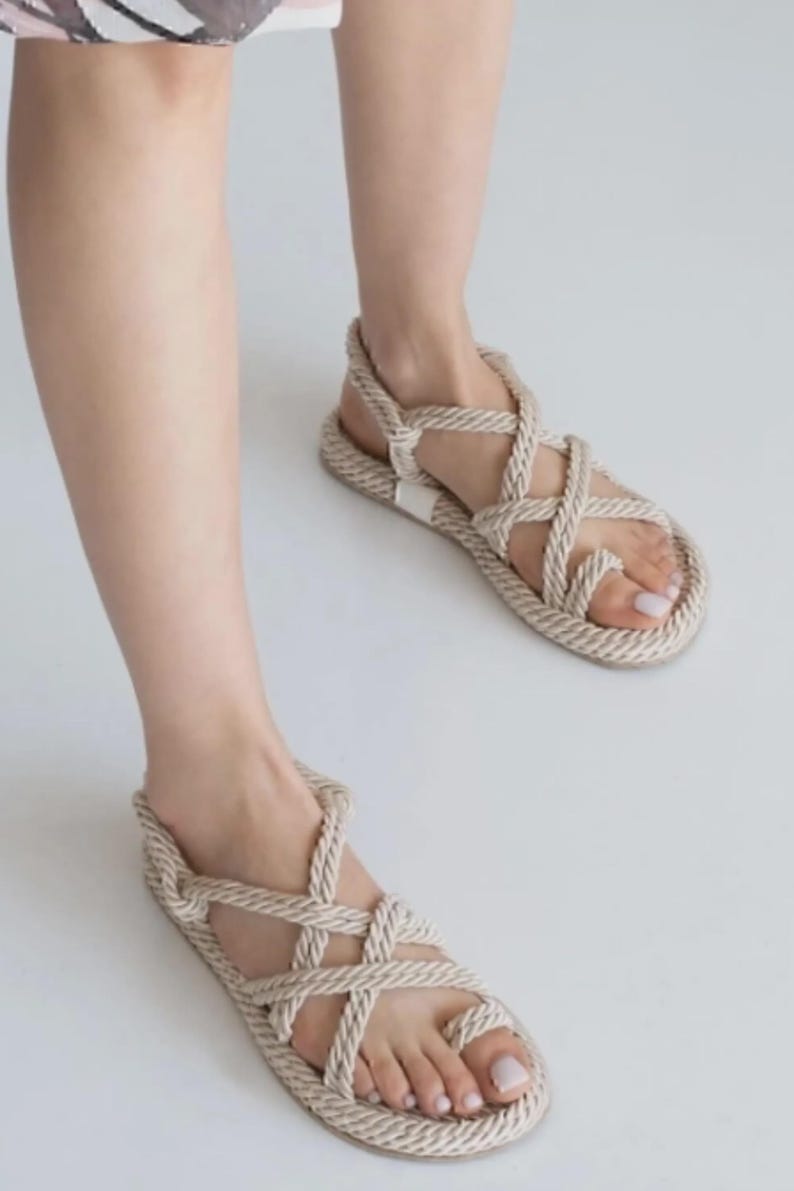 May include: Beige rope sandals with a toe loop and crisscross straps. The sandals have a flat sole and a strap around the ankle. The footwear is displayed on a white background.
