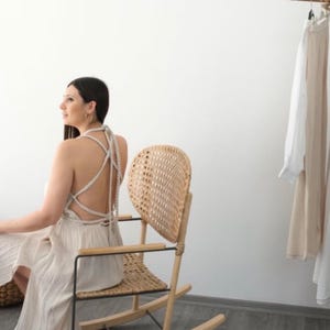 May include: A woman wearing a white crochet dress with a criss-cross back detail sits in a wicker rocking chair. The chair has a unique heart-shaped back.
