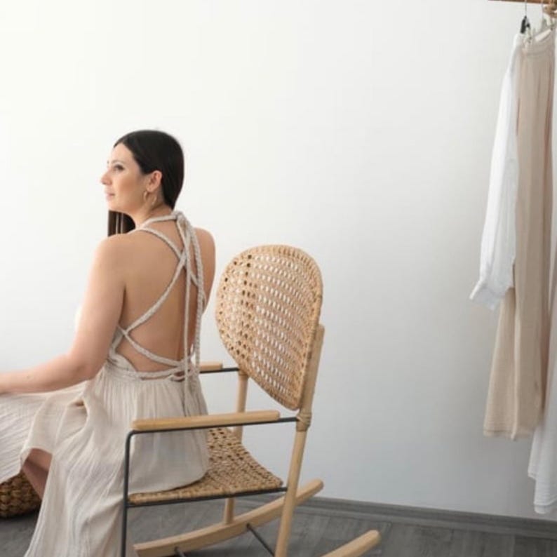 May include: A woman wearing a white crochet dress with a criss-cross back detail sits in a wicker rocking chair. The chair has a unique heart-shaped back.
