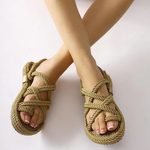 May include: Tan rope sandals with a woven design. The sandals feature a toe loop and ankle strap, and are shown on a white background. The sandals appear to be made of a thick, durable rope material.