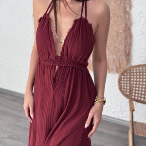 May include: A burgundy halter maxi dress with a deep V-neckline and a high slit. The dress features a textured fabric and a gathered waist. A gold necklace and bracelets are visible.
