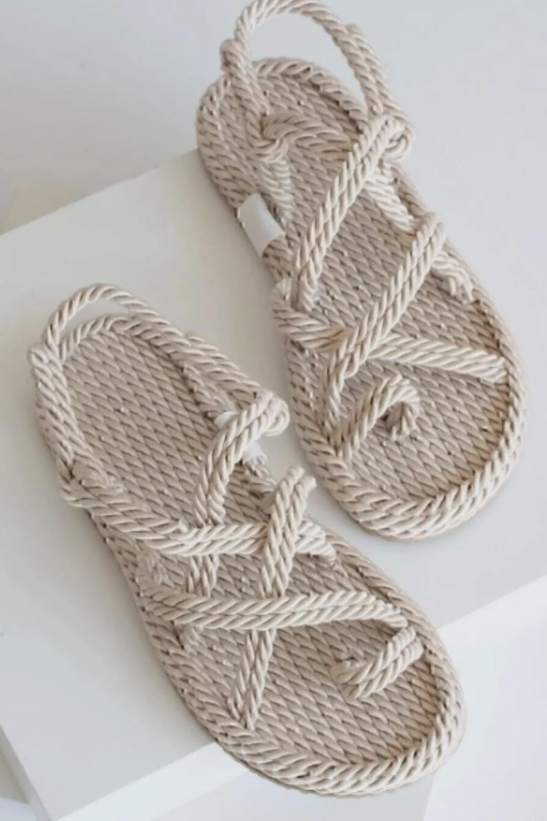May include: A pair of beige rope sandals. The sandals feature a woven design with a toe loop and straps that crisscross over the foot. The soles are flat and the sandals appear to be made of a natural fiber.