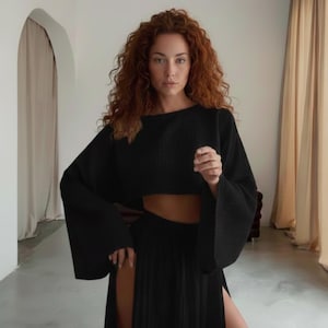 May include: A black two-piece outfit featuring a cropped top with wide sleeves and a high-slit skirt. The outfit is made of a textured fabric. The model has long, curly red hair.