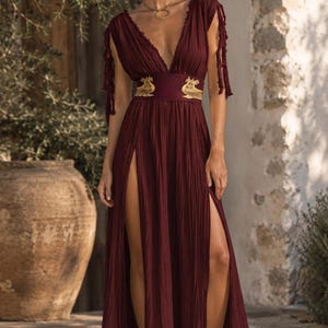 May include: A floor-length, deep burgundy dress featuring a deep V-neckline, short sleeves with fringe detailing, and two high side slits. The waist is cinched with a wide band adorned with gold-coloured embroidered animal motifs.