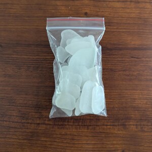 May include: A transparent plastic bag containing smooth, frosted, white sea glass pieces. The sea glass has rounded edges and various shapes. The bag is sealed with a red zip and rests on a dark brown wooden surface.