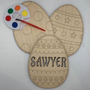 Pode incluir: Três ovos de Páscoa de madeira inacabados com tinta e um pincel. Os ovos são decorados com diferentes padrões, incluindo círculos, estrelas e linhas onduladas. Um ovo tem o nome "Sawyer" escrito nele.