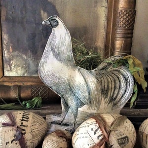 May include: A black and white illustration of a rooster with a long tail, printed on a paper cutout. The rooster is standing on a wooden surface with five decorative eggs made of paper and wrapped in newspaper.