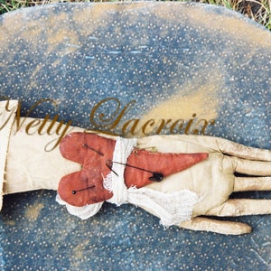 May include: A red felt heart with safety pins and white fabric is tied around a white fabric glove on a blue and white patterned surface. The text "Netty Lacroix" is written in cursive on the surface.