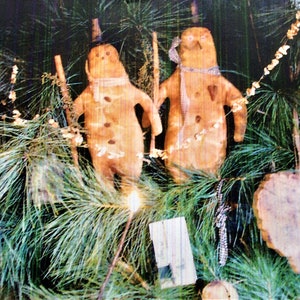 May include: Two rustic, brown, snowman-shaped ornaments with white accents are hanging on a green pine tree branch. The ornaments have a simple, handmade look and are decorated with buttons and a heart.