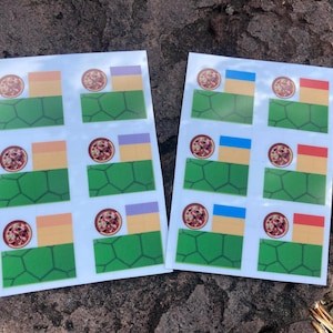 May include: Two sheets of stickers with a pizza graphic and colorful geometric designs. Each sticker features a pizza, a green polygon, and a rectangle in various colors like orange, blue, and purple. The stickers are on a textured, brown surface.
