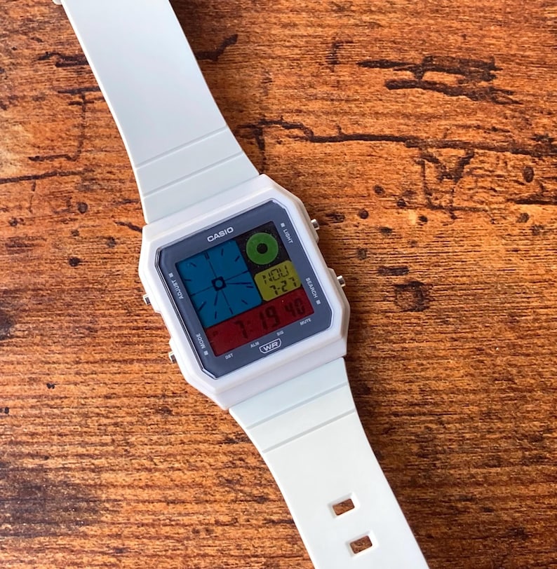 May include: A white Casio digital watch with a multi-coloured display. The watch face has a digital display showing the time, date, and day of the week. The watch has a white strap and a white case.