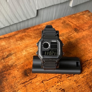 Pu&ograve; includere: Un orologio digitale Casio nero con quadrante quadrato e cinturino nero. L'orologio ha un display digitale che mostra l'ora come 11:02. L'orologio &egrave; su un supporto nero.