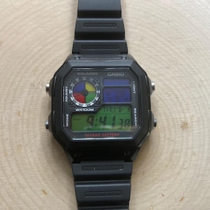 May include: Black Casio digital watch with a rectangular face and black strap. The watch face shows the time, day, and date in green, blue, and multi-coloured sections. The watch has buttons for adjusting the mode, light, and search functions.