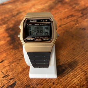 May include: A gold Casio F-91W digital watch with a black band. The watch face displays the time, date, and day of the week. The watch is water resistant.