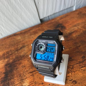 Pu&ograve; includere: Orologio digitale Casio nero con un display digitale blu che mostra l'ora 5:58:29. L'orologio ha un cinturino in resina nera e una cassa in resina nera. L'orologio ha una funzione di ora mondiale, una batteria da 10 anni e un illuminatore.