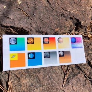 May include: A white rectangular sticker sheet featuring colorful illustrations of planets against vibrant backgrounds. The planets include Earth, Venus, Mars, Jupiter, and others, each set within a square. The sticker sheet is placed on a rough, brown rock surface.
