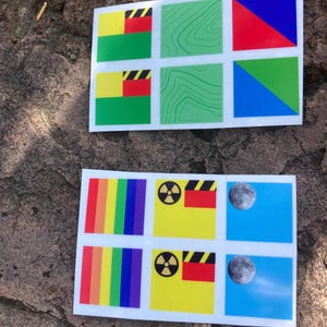 May include: Two sheets of stickers with various designs. One sheet features colorful geometric shapes and patterns, including a rainbow, a nuclear symbol, and a moon. The other sheet has flags and topographic designs. The stickers are on a stone surface.