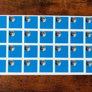 Puede incluir: Una hoja de pegatinas azules con borde blanco, cada una con una imagen en blanco y negro de una luna creciente.