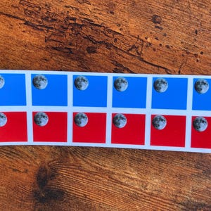 May include: A sticker sheet featuring twelve moon phase stickers. Each sticker has a detailed image of the moon against a blue or red background. The stickers are arranged in a grid pattern.