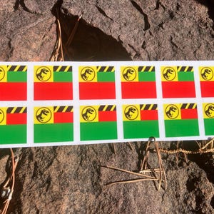 May include: A sheet of stickers featuring a logo with a dinosaur silhouette inside a yellow circle, set against a red, green, and black striped background. The stickers are rectangular and arranged in rows.
