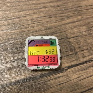 May include: A small, square digital watch with a white frame. The display shows "NYC 3:32" and "1:32:38" in red and yellow. The top of the watch has a colorful graphic of a train and a palm tree.