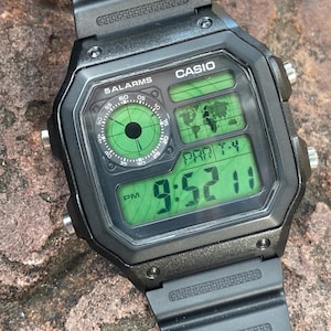 May include: Black Casio digital watch with a square face and a black strap. The watch face displays the time, date, and other functions in green digital numbers. The watch has the text "CASIO" and "5 ALARMS" on the face.