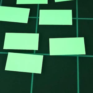 May include: A dark green cutting mat with a grid pattern and measurement markings. Several small, rectangular, light green stickers are scattered across the surface, near the 8 and 9 inch markings.