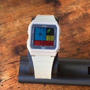 May include: A white Casio digital wristwatch with a black face and a multi-coloured display. The watch has a black band and a silver buckle. The display shows the time, date, and day of the week.