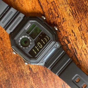 Pu&ograve; includere: Un orologio digitale Casio nero con quadrante quadrato e cinturino in resina nera. Il quadrante dell'orologio mostra l'ora, la data e il giorno della settimana. L'orologio ha una retroilluminazione verde.