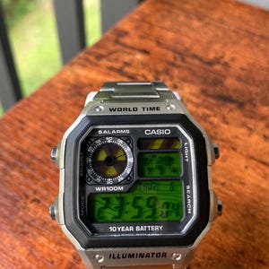 Pode incluir: Relógio digital Casio prateado com mostrador preto e pulseira de aço inoxidável. O relógio exibe a hora, a data e outras funções em uma tela digital verde. Possui hora mundial, alarmes e bateria de 10 anos.