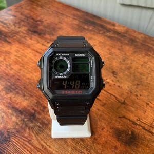 Op de afbeelding: Een zwarte digitale Casio-horloge met een vierkante wijzerplaat, die de tijd 4:48:00 weergeeft. Het horloge heeft meerdere digitale displays en knoppen en staat op een witte standaard. De horlogeband is ook zwart.