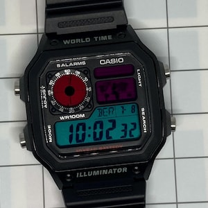 May include: Black digital wristwatch with a red and black compass dial, a digital display showing the time as 10:02:32, and the text "WORLD TIME", "SALARMS", "CASIO", "LIGHT", "SEARCH", "WR100M", "BER 1 8", and "ILLUMINATOR".