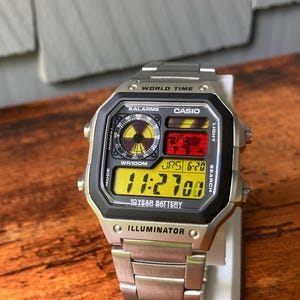 Puede incluir: Un reloj digital Casio plateado con un bisel negro y una pantalla digital amarilla. La esfera del reloj muestra la hora, la fecha y las funciones de hora mundial. El reloj tiene una correa de acero inoxidable y el texto "WORLD TIME", "CASIO" e "ILLUMINATOR" son visibles.