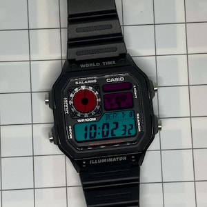 May include: Black Casio digital watch with a rectangular face. The watch displays the time, day, and date in a teal-colored digital display. The watch has a black band and features the text "CASIO" and "WORLD TIME".