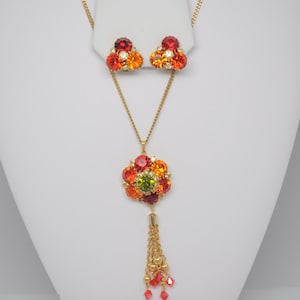 May include: A gold necklace with a pendant featuring a cluster of red, orange, and green rhinestones. The pendant has a gold chain tassel. Matching earrings with red, orange, and clear rhinestones are also included.