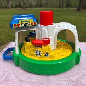 May include: A colorful toy train station with a yellow train on a circular track. The station has a blue roof with the text "SHELCORE" and a red cylinder. The base is green, and the track is yellow.