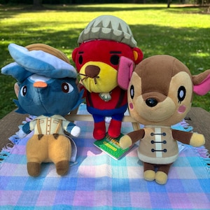 May include: Three plush toys of cartoon characters are displayed on a checkered blanket. The toys include a blue penguin, a red bear, and a brown deer. The penguin wears a hat and overalls. The bear wears a hat and shorts. The deer wears a jacket.