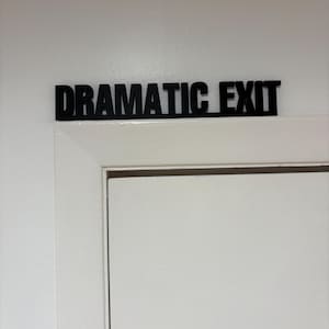 May include: Black sign that reads "DRAMATIC EXIT" mounted above a white door. The sign is in a bold, sans-serif font. The door and surrounding wall are painted white.