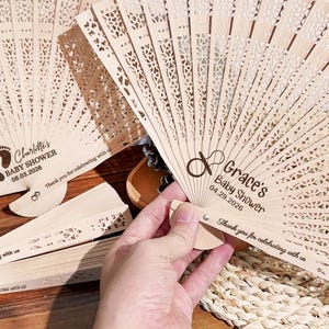 May include: Several intricately carved wooden fans. One is held, showing the text "Grace's Baby Shower 04.29.2026". Another fan reads "Charlotte's Baby Shower 08.03.2026". Both fans have the phrase "Thank you for celebrating with us".