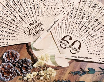 Op de afbeelding: Twee ingewikkeld gesneden houten waaiers. Op één waaier staat "mis quince años" in script. De andere waaier leest "60 Happy Birthday!" met de naam "Melissa" eronder. De waaiers liggen op een houten oppervlak met dennenappels en bloemen.