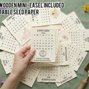 May include: A collection of 2026 calendars made from plantable seed paper. Each calendar page features a month, with the month's name printed at the top. The calendars are beige with floral and bee illustrations. The text "FREE WOODEN MINI-EASEL INCLUDED PLANTABLE SEED PAPER" is at the top.